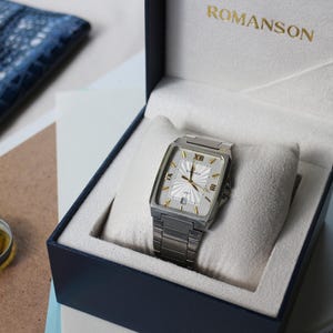 May include: A rectangular silver-tone wristwatch with a white dial, gold-tone indices, and a date window, resting on a white cushion inside a navy blue presentation box with the gold text ROMANSON printed on the interior lid.