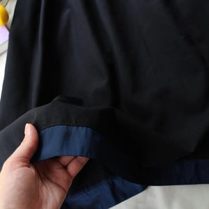 K&ouml;nnte beinhalten: Nahaufnahme eines schwarzen Stoffes mit marineblauer Paspel. Der Stoff wird hochgehalten und zeigt die Kontrastfarbe. Das Bild ist gut beleuchtet und hebt die Textur und Farbe des Materials hervor. Die Gesamt&auml;sthetik ist schlicht und elegant.