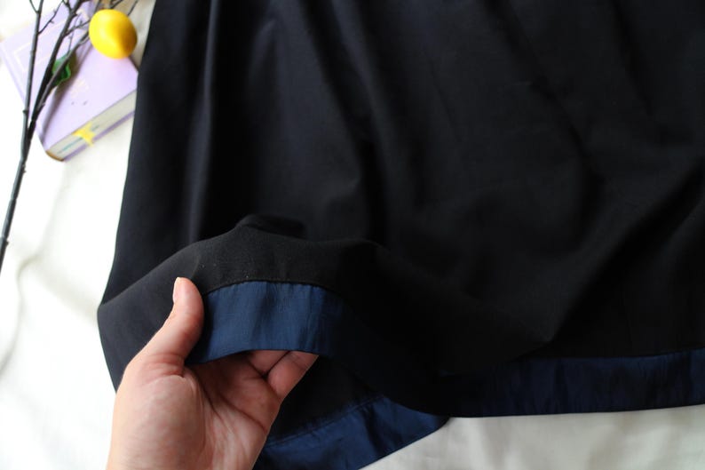 K&ouml;nnte beinhalten: Nahaufnahme eines schwarzen Stoffes mit marineblauer Paspel. Der Stoff wird hochgehalten und zeigt die Kontrastfarbe. Das Bild ist gut beleuchtet und hebt die Textur und Farbe des Materials hervor. Die Gesamt&auml;sthetik ist schlicht und elegant.