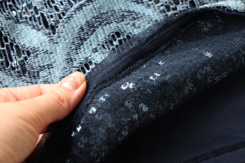 May include: Close-up of a dark blue fabric with a layered design. The top layer is a delicate, open-weave lace, while the bottom layer appears to be a solid, opaque material. The edges are neatly sewn together.