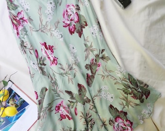 Puede incluir: Una falda midi de color verde claro con un estampado floral de rosas rosa intenso, hojas verde oscuro y pequeñas flores blancas. La falda presenta un dobladillo acampanado y una confección en tejido ligero y liso con estampado botánico.