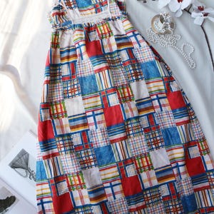 May include: A vintage patchwork dress with a square pattern in red, blue, yellow, and white. The dress has a scoop neckline, ruffled sleeves, and a long skirt. The dress is made of a lightweight fabric.