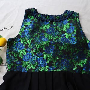 K&ouml;nnte beinhalten: &Auml;rmelloses Kleid mit schwarzem Faltenrock und floralem Oberteil. Das Oberteil hat einen dunklen Hintergrund mit blauen und gr&uuml;nen Blumenmustern. Der Ausschnitt ist mit einem R&uuml;schendetail verziert. Das Kleid wird auf einer wei&szlig;en Oberfl&auml;che pr&auml;sentiert.