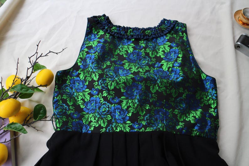 K&ouml;nnte beinhalten: &Auml;rmelloses Kleid mit schwarzem Faltenrock und floralem Oberteil. Das Oberteil hat einen dunklen Hintergrund mit blauen und gr&uuml;nen Blumenmustern. Der Ausschnitt ist mit einem R&uuml;schendetail verziert. Das Kleid wird auf einer wei&szlig;en Oberfl&auml;che pr&auml;sentiert.