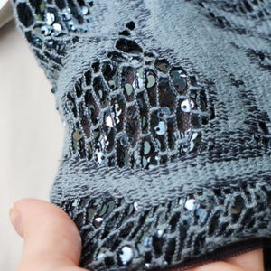 May include: Close-up of a section of a blue-gray fabric with a net-like design. The fabric is embellished with small, shiny, dark blue sequins. The net pattern reveals a darker fabric underneath, creating a textured effect.