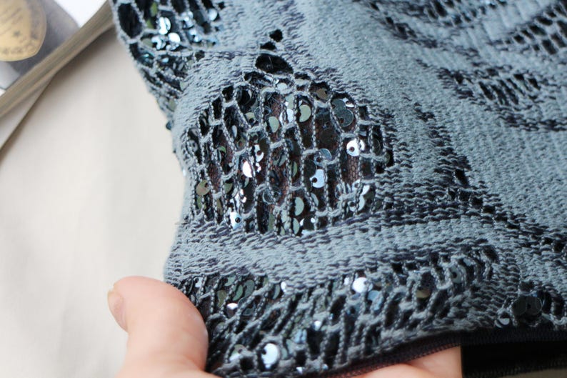 May include: Close-up of a section of a blue-gray fabric with a net-like design. The fabric is embellished with small, shiny, dark blue sequins. The net pattern reveals a darker fabric underneath, creating a textured effect.