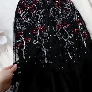 May include: Black evening gown with intricate sequin and bead embellishments. The design features floral patterns in red and silver, creating a shimmering effect. The gown is made of a sheer fabric, with a flowing silhouette.