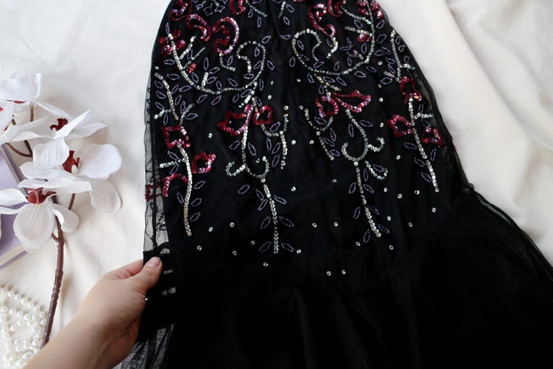 May include: Black evening gown with intricate sequin and bead embellishments. The design features floral patterns in red and silver, creating a shimmering effect. The gown is made of a sheer fabric, with a flowing silhouette.