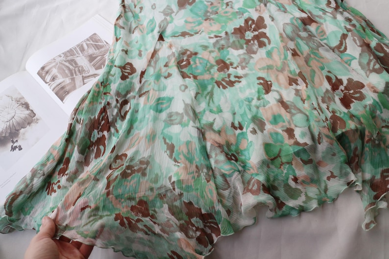 May include: A flowing, sheer fabric with a floral pattern in shades of green, brown, and peach. The fabric is draped over an open book, with the edges of the fabric slightly ruffled. The overall effect is soft and delicate.