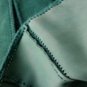 May include: Close-up of teal-colored leather pieces stitched together. The edges are meticulously sewn with a contrasting thread, creating a textured, geometric pattern. The leather has a soft, matte finish, and the stitching adds a decorative element.