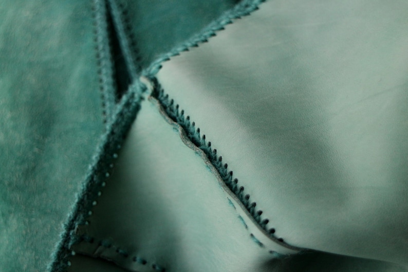 May include: Close-up of teal-colored leather pieces stitched together. The edges are meticulously sewn with a contrasting thread, creating a textured, geometric pattern. The leather has a soft, matte finish, and the stitching adds a decorative element.