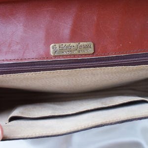 May include: Open view of a brown leather handbag interior. The bag features a zippered pocket, a small open pocket, and a light tan lining. A gold-toned metal tag reads "Catalan Garces Made in Spain."