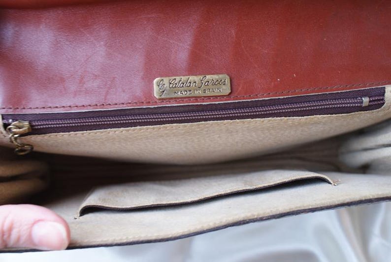 May include: Open view of a brown leather handbag interior. The bag features a zippered pocket, a small open pocket, and a light tan lining. A gold-toned metal tag reads "Catalan Garces Made in Spain."
