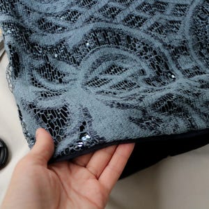 May include: Close-up of a section of a garment with a gray lace overlay. The lace features a swirling floral pattern and is embellished with small sequins. The garment has a dark lining and is being held by a hand.