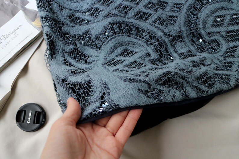 May include: Close-up of a section of a garment with a gray lace overlay. The lace features a swirling floral pattern and is embellished with small sequins. The garment has a dark lining and is being held by a hand.