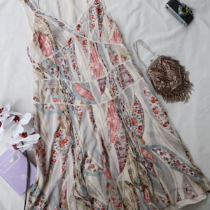 May include: A sleeveless, V-neck dress with a floral and leaf pattern in shades of pink, red, and blue on a cream background. The dress has a tiered design with lace trim. A small beaded purse and a vintage camera are also visible.