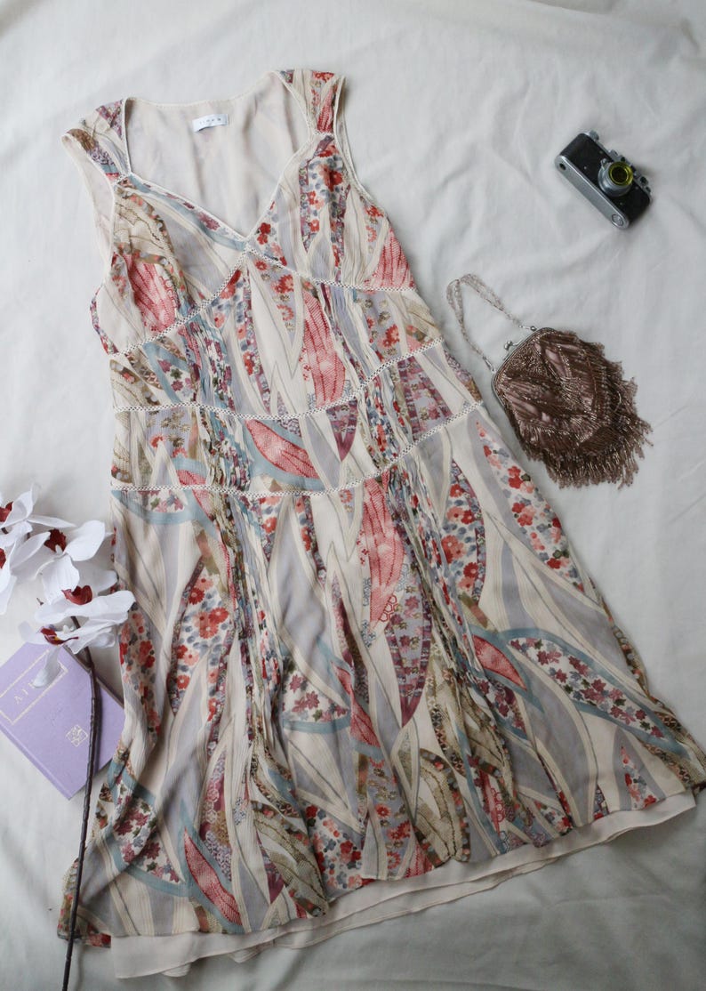 May include: A sleeveless, V-neck dress with a floral and leaf pattern in shades of pink, red, and blue on a cream background. The dress has a tiered design with lace trim. A small beaded purse and a vintage camera are also visible.