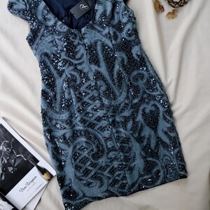 May include: A sleeveless, knee-length dress in a muted blue hue, adorned with intricate sequined patterns. The dress features a scoop neckline and a dark blue lining. Accessories include a braided bracelet and gold-toned earrings.