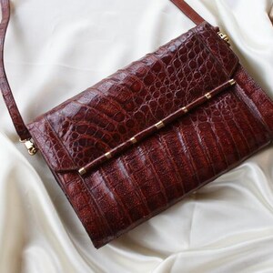 May include: A brown leather clutch with a textured, crocodile-like pattern. The bag has a long, thin strap and a gold-colored bar with small gold accents. The clutch is on a white satin surface.