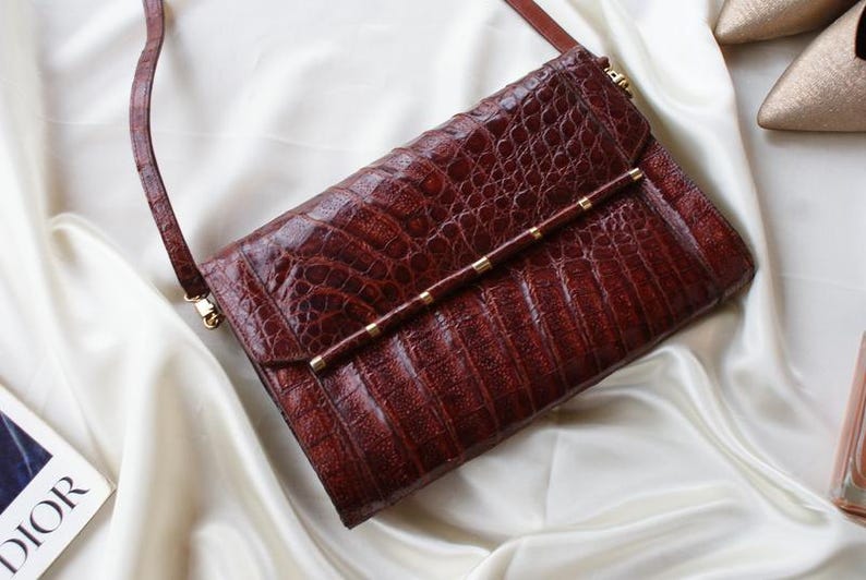 May include: A brown leather clutch with a textured, crocodile-like pattern. The bag has a long, thin strap and a gold-colored bar with small gold accents. The clutch is on a white satin surface.