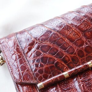 May include: A close-up of a brown leather handbag with a crocodile skin texture. The bag has a long strap and a gold-colored clasp. The bag's surface is a rich, reddish-brown color.