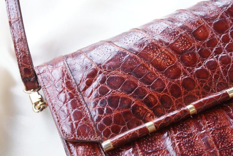 May include: A close-up of a brown leather handbag with a crocodile skin texture. The bag has a long strap and a gold-colored clasp. The bag's surface is a rich, reddish-brown color.