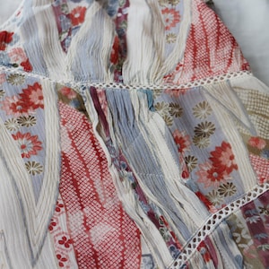 May include: Close-up of a patchwork fabric with floral and geometric patterns. The fabric features a mix of red, blue, and white hues, with delicate lace trim. The design includes stripes and floral motifs, creating a textured, layered effect.