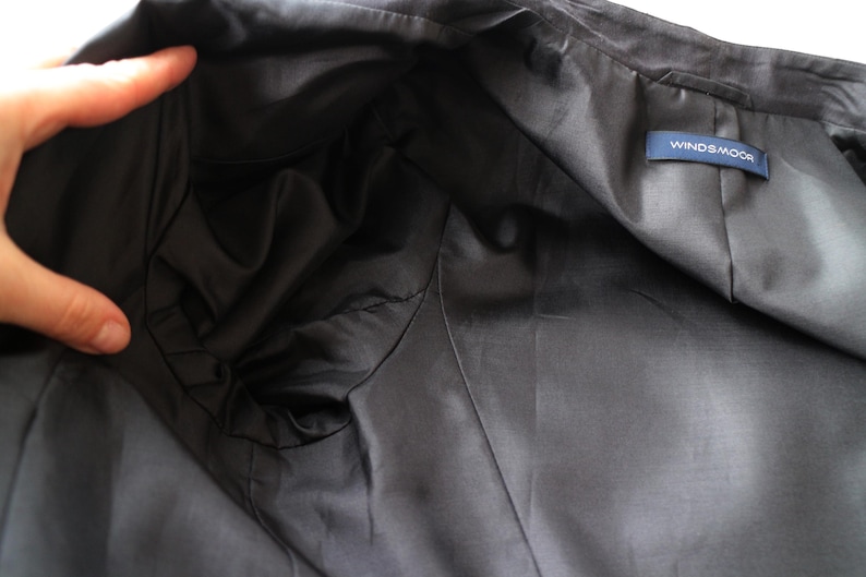 May include: Close-up of the inside of a dark gray jacket. The lining is a darker shade of gray and appears smooth. A small blue tag with the text "WINDSMOOR" is visible.