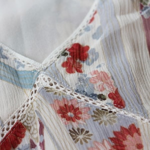May include: Close-up of a floral patterned fabric. The fabric features a white base with vertical stripes and a delicate floral print in shades of red, pink, blue, and brown. The edges are trimmed with white lace.