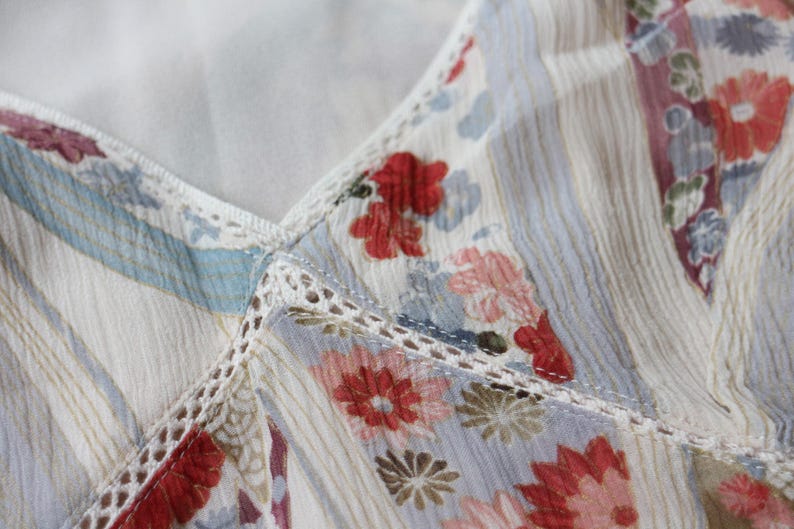 May include: Close-up of a floral patterned fabric. The fabric features a white base with vertical stripes and a delicate floral print in shades of red, pink, blue, and brown. The edges are trimmed with white lace.