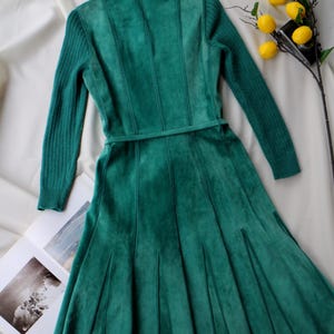 May include: A teal green dress with long sleeves and a fitted bodice. The dress features a flared skirt and a matching belt. The sleeves are ribbed, and the bodice has vertical seams. A sprig of lemons sits nearby.