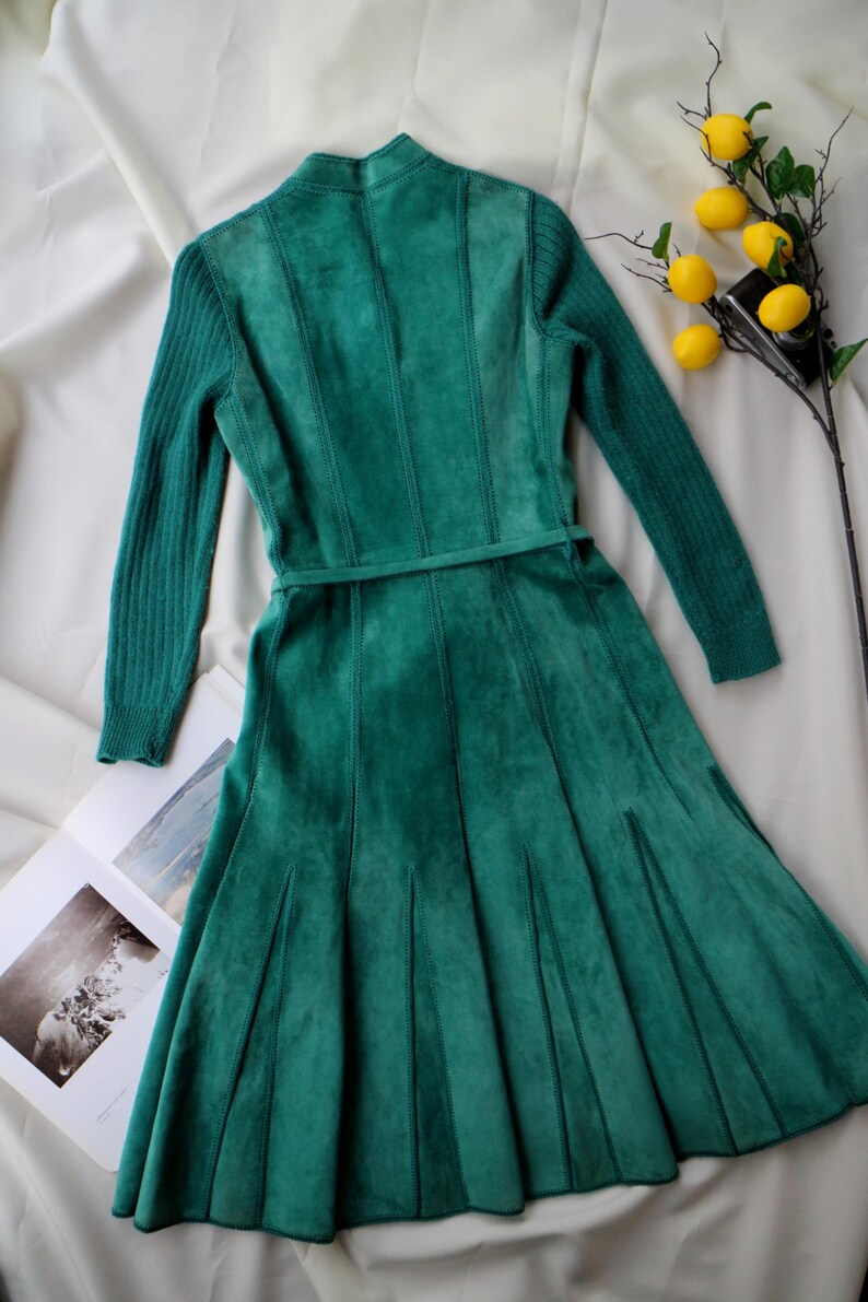 May include: A teal green dress with long sleeves and a fitted bodice. The dress features a flared skirt and a matching belt. The sleeves are ribbed, and the bodice has vertical seams. A sprig of lemons sits nearby.