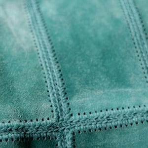 May include: Close-up of a teal suede fabric with a stitched, patchwork design. The stitching is a darker teal color, creating a textured, geometric pattern. The suede has a soft, slightly worn appearance.