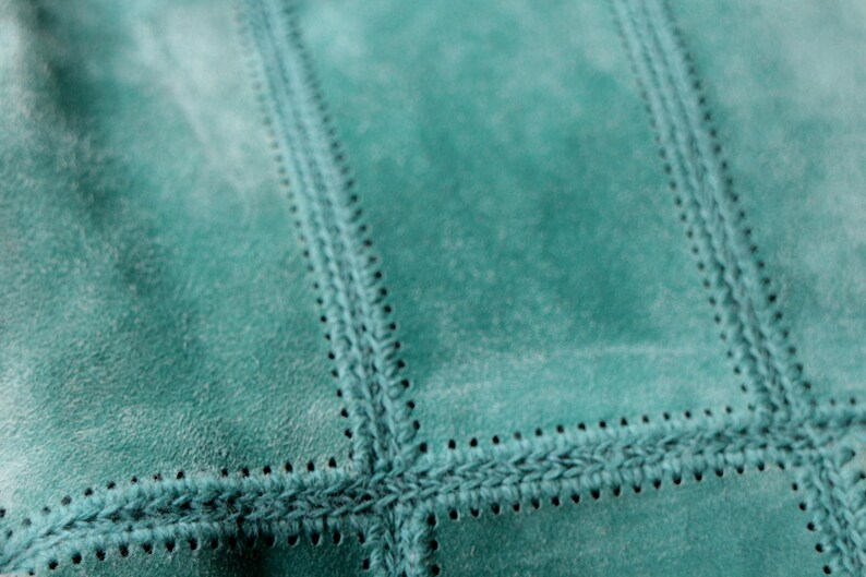 May include: Close-up of a teal suede fabric with a stitched, patchwork design. The stitching is a darker teal color, creating a textured, geometric pattern. The suede has a soft, slightly worn appearance.