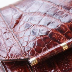 May include: Close-up of a brown leather clutch with a crocodile skin texture. The clutch has a quilted pattern and a gold-colored clasp. The leather appears to be a deep reddish-brown color.