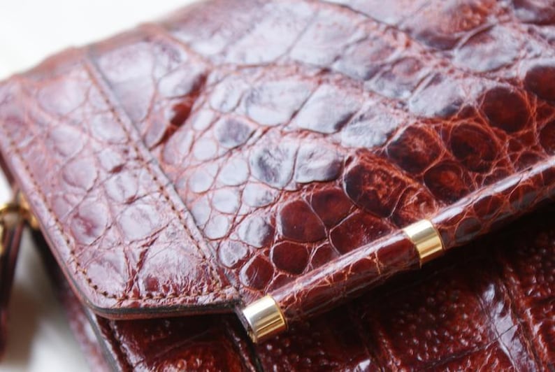 May include: Close-up of a brown leather clutch with a crocodile skin texture. The clutch has a quilted pattern and a gold-colored clasp. The leather appears to be a deep reddish-brown color.