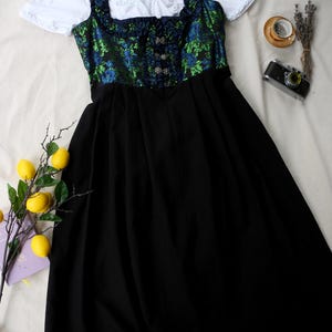 K&ouml;nnte beinhalten: Ein traditionelles deutsches Dirndl-Kleid. Das Oberteil hat ein gr&uuml;nes und blaues Blumenmuster, mit einem schwarzen Rock und einer wei&szlig;en Bluse. Das Kleid hat dekorative Kn&ouml;pfe und eine taillierte Silhouette. Das Kleid wird auf einer wei&szlig;en Oberfl&auml;che pr&auml;sentiert.