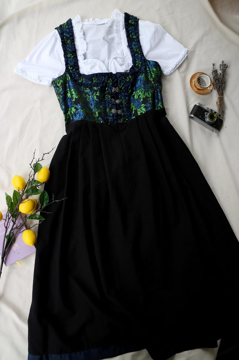 K&ouml;nnte beinhalten: Ein traditionelles deutsches Dirndl-Kleid. Das Oberteil hat ein gr&uuml;nes und blaues Blumenmuster, mit einem schwarzen Rock und einer wei&szlig;en Bluse. Das Kleid hat dekorative Kn&ouml;pfe und eine taillierte Silhouette. Das Kleid wird auf einer wei&szlig;en Oberfl&auml;che pr&auml;sentiert.
