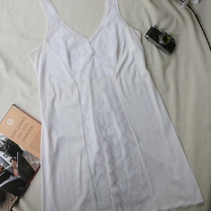 May include: A white slip dress featuring a V-neck and intricate lace paneling down the front. The dress has a sleeveless design and is made from a soft material. A vintage camera and a magazine are also visible.