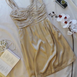May include: A champagne-colored satin dress with a ruched bodice and spaghetti straps. The dress is sleeveless and has a knee-length skirt. A small vintage camera, a sprig of white orchids, and a book are next to the dress.