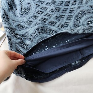 May include: A close-up of a blue sequined top with a patterned lace overlay. The top has a dark blue lining and is partially held open, revealing the interior. The fabric appears soft and delicate.