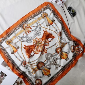 May include: Square silk scarf with a horse design. The scarf has a white background with brown horse heads, ribbons, and chains. It has an orange border and decorative elements.