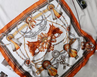 May include: Square silk scarf with a horse-themed design. The scarf features a white background with brown horse heads, ribbons, and chains. It has an orange border and decorative elements.
