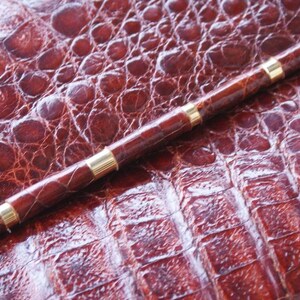 May include: A brown pen with gold-colored bands rests on a textured, reddish-brown surface. The pen's body is a darker shade of brown, and the gold bands add a touch of elegance. The surface has a reptile-like pattern.