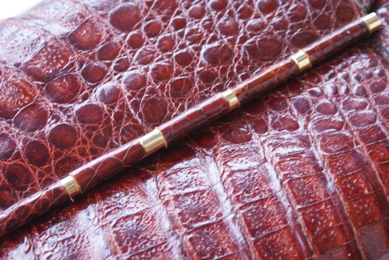 May include: A brown pen with gold-colored bands rests on a textured, reddish-brown surface. The pen's body is a darker shade of brown, and the gold bands add a touch of elegance. The surface has a reptile-like pattern.