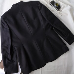 May include: A black blazer with a tailored fit, featuring a classic design. The jacket is displayed on a white fabric background, with a vintage camera and pearl necklace adding a touch of elegance. The blazer has long sleeves.