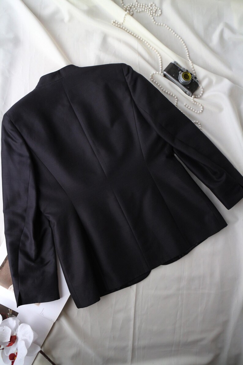 May include: A black blazer with a tailored fit, featuring a classic design. The jacket is displayed on a white fabric background, with a vintage camera and pearl necklace adding a touch of elegance. The blazer has long sleeves.
