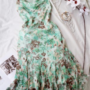 May include: A flowing, mint green dress with a floral pattern in shades of brown and green. The dress has a cowl neckline and a layered skirt. A pearl necklace, a small pendant, and a white orchid are also visible.