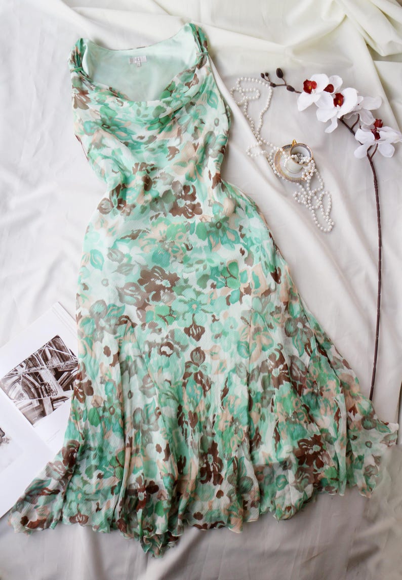May include: A flowing, mint green dress with a floral pattern in shades of brown and green. The dress has a cowl neckline and a layered skirt. A pearl necklace, a small pendant, and a white orchid are also visible.