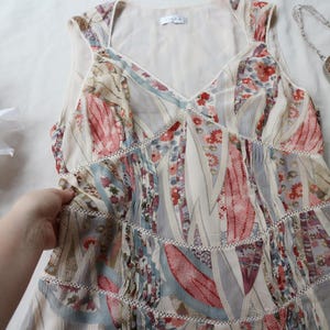 May include: A sleeveless, floral print dress with a V-neckline and tiered skirt. The dress features a mix of red, blue, and cream-colored patterns. A small, beaded purse with fringe is visible in the background.